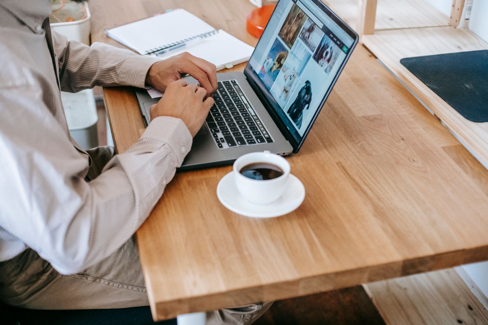 Person in a striped shirt typing on a laptop at a wooden desk, with a notebook and a cup of coffee beside it