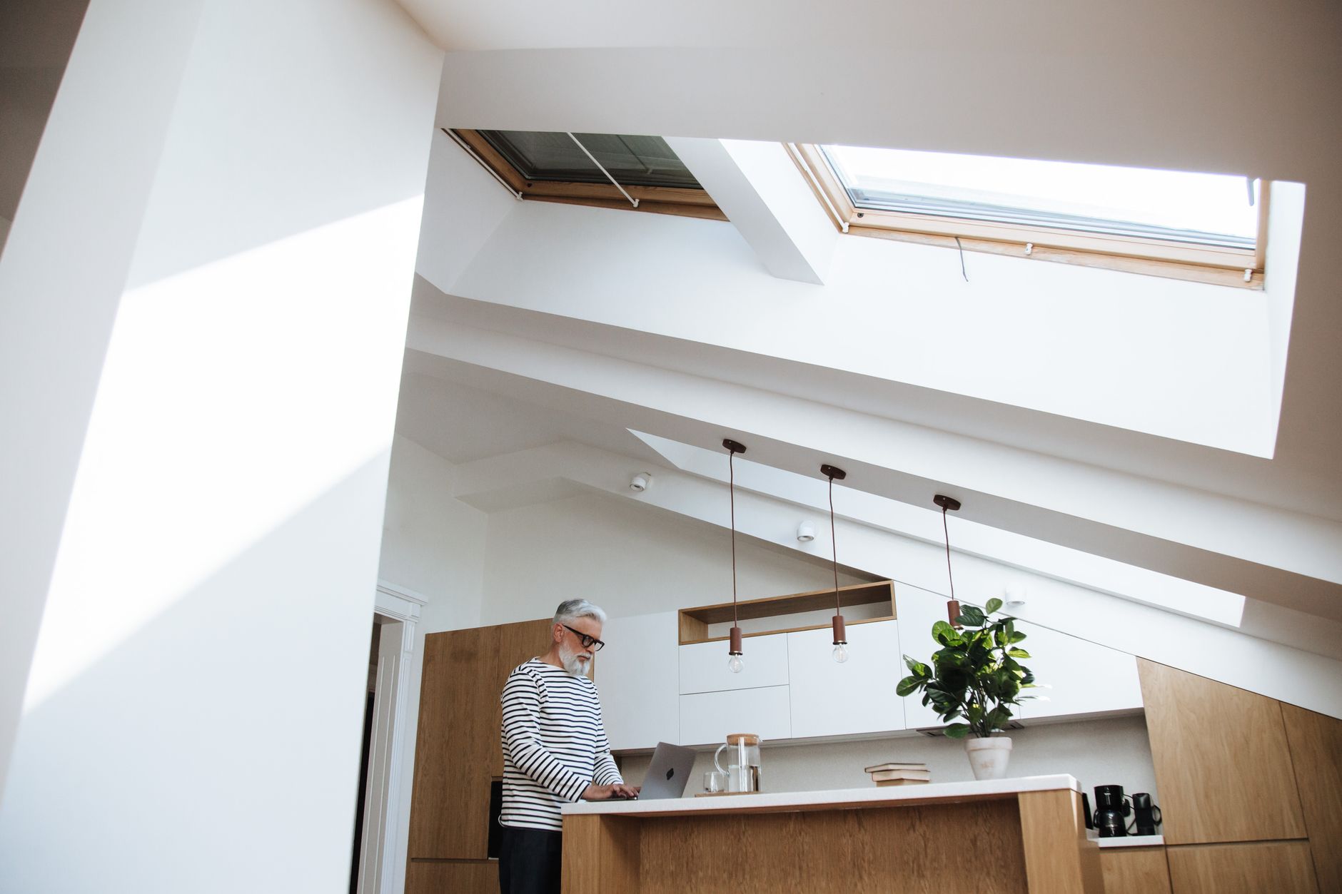 Gray-haired man in a striped top working at a laptop on a kitchen counter in a bright home with skylights